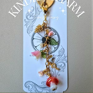 May include: A gold-coloured Kindle charm featuring a heart-shaped clasp, decorated with small flowers, a mushroom, and a key charm. The charm is suspended from a decorative tag with the text "Kindle Charm" and "Forest Fairy".