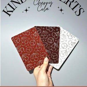 May include: Three Kindle inserts with cherry cola designs. The inserts feature a pattern of cherries and ice cubes in red, dark brown, and white. The text "KINDLE INSERTS Cherry Cola" is arched above the inserts.