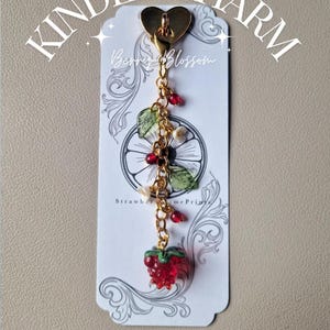 May include: A gold-toned Kindle charm featuring a heart-shaped attachment, a chain with small red beads, white flower accents, green leaves, and a red strawberry pendant. The charm is displayed on a white card with the text Kindle Charm, Berry & Blossom, StrawberryLimePrints, Sticky Hook Included, and Easily Detachable.