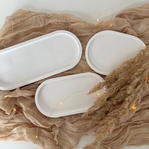 May include: Three white, oval-shaped serving trays of varying sizes are arranged on a beige, textured fabric. Dried, tan-colored ornamental grass and a string of tiny lights add decorative elements to the composition. The trays are smooth and minimalist.