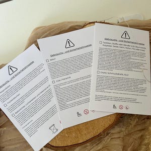 Può includere: Tre carte bianche con testo tedesco e un simbolo di avvertimento sono disposte su una fetta di legno e tessuto beige. Le carte contengono informazioni sull'uso e la sicurezza del prodotto. Le carte sono per articoli decorativi.