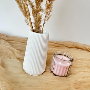 Puede incluir: Un jarrón de cerámica blanco y acanalado con hierba de la pampa seca, junto a una pequeña vela de tarro de cristal con cera rosa. La vela está en un tarro de cristal transparente con un diseño acanalado.