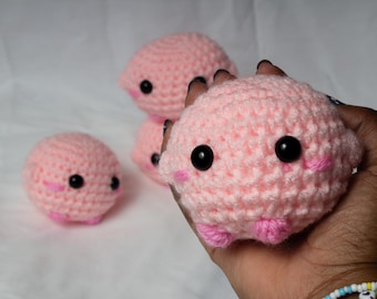 May include: Handmade crochet plushies in soft pink. Each plushie features black safety eyes and pink accents for cheeks and feet. The image shows several of these adorable, round, and cuddly toys.
