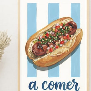 Pode incluir: Uma impressão emoldurada com um cachorro-quente num pão, coberto com vegetais e ervas picadas. O fundo tem listras azuis e brancas. As palavras "a comer" estão escritas em azul na parte inferior.