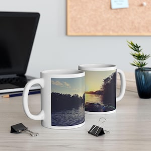 Pode incluir: Duas canecas de cerâmica branca com uma imagem panorâmica de um lago e árvores ao pôr do sol. A imagem na caneca à esquerda mostra uma visão mais próxima do lago e das árvores. A caneca à direita mostra uma visão mais ampla do lago e das árvores.