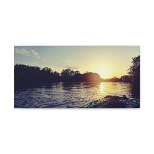 以下が含まれることがあります： 遠くで沈む夕日を背景に、湖畔に木々がシルエットを描いています。太陽は明るく輝き、水面に金色の光を投げかけています。手前に、一部が写っているカヤックがあります。