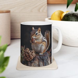 May include: A white ceramic mug with a brown and black image of a chipmunk eating an acorn. The chipmunk is sitting on a tree stump with a pine cone in the background. The mug has a black background and a white handle.