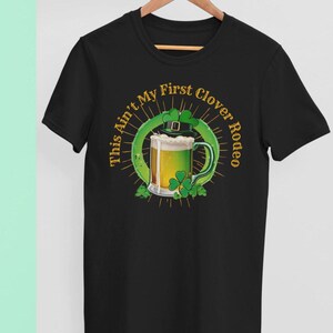 May include: Black t-shirt with a green shamrock and a glass of beer. The text on the shirt reads "This Ain't My First Clover Rodeo".