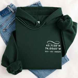 May include: A dark green hoodie with a drawstring hood and a white embroidered design. The design includes coordinates and Roman numerals. The hoodie is folded on top of a pair of blue jeans.