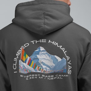 Puede incluir: Sudadera con capucha gris oscuro con un gráfico en la espalda que dice "I Climbed the Himalayas" en un arco blanco. El gráfico incluye una ilustración de montaña, banderas de oración y texto que indica el campamento base del Everest a 5.364 metros en Nepal.