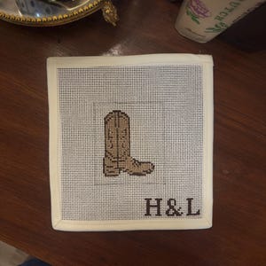 May include: Square, off-white needlepoint coaster featuring a brown cowboy boot design. The initials "H&L" are stitched in black below the boot. The coaster has a cream-colored border and is set on a wooden surface.