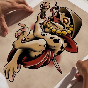 Puede incluir: Ilustraci&oacute;n estilo tatuaje de una criatura parecida a una rana con una daga, que lleva una capa roja y un collar de cuentas doradas. La criatura tiene una capucha marr&oacute;n y est&aacute; sobre un fondo beige. La imagen est&aacute; sujeta por dos manos.