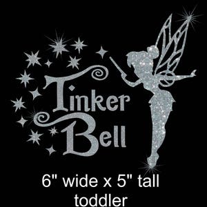 May include: Silver glitter iron-on design featuring Tinker Bell with a wand and stars, with the text "Tinker Bell" in a script font. The design is 6 inches wide and 5 inches tall, suitable for a toddler.