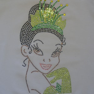 May include: A rhinestone design of a cartoon princess with a green and yellow crown. The princess is smiling and has brown eyes.