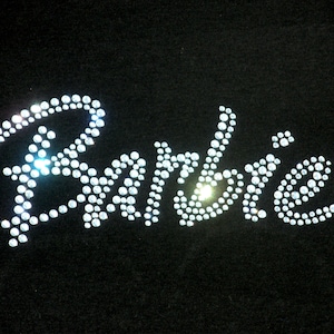 May include: The word Barbie is written in a cursive script font on a solid black fabric background. The lettering is formed entirely by small, clear, reflective rhinestones that catch the light, creating a sparkling effect against the dark surface.