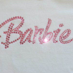 May include: A white fabric surface featuring the word Barbie written in a cursive script. The lettering is formed by numerous small, round, pink rhinestones that are arranged to create the text, with a bright light reflection on the letter b.