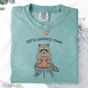 May include: Seafoam green Comfort Colors t-shirt with the text "Off to pottery class" above a cartoon raccoon at a pottery wheel. The raccoon is working with a clay pot. A silver necklace with a pendant is on the shirt.