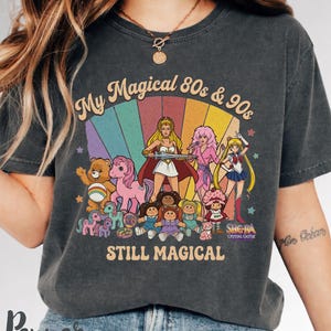 My Magical 80s & 90s" graphic on a charcoal grey Comfort Colors 1717 t-shirt. The vintage-style design features a collage of nostalgic characters including She-Ra, Jem, Sailor Moon, Care Bears, My Little Pony, Cabbage Patch Kids, and Strawberry Shortcake against a retro rainbow backdrop. The text reads "Still Magical" in a classic 80s font. Perfect for fans of retro-futurism, cottagecore, and nostalgic pop culture fashion.
