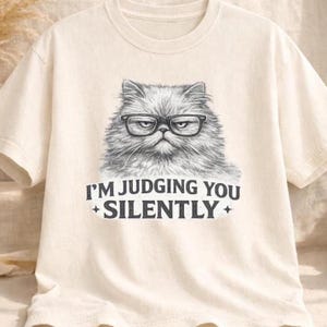 Może przedstawiać: Kremowa koszulka z szczegółową ilustracją kota w okularach. Kot ma surowy wyraz twarzy. Poniżej napis "I'M JUDGING YOU SILENTLY" z gwiazdkami.