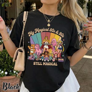May include: Black t-shirt with a colorful graphic featuring cartoon characters from the 80s and 90s, including Care Bears, My Little Pony, and Sailor Moon. The text on the shirt reads "My Magical 80s & 90s STILL MAGICAL."