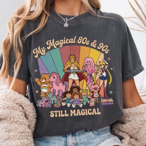 May include: Dark gray t-shirt with a colorful graphic featuring characters from the 80s and 90s, including She-Ra, Sailor Moon, Care Bears, and My Little Pony. The text reads "My Magical 80s & 90s STILL MAGICAL".