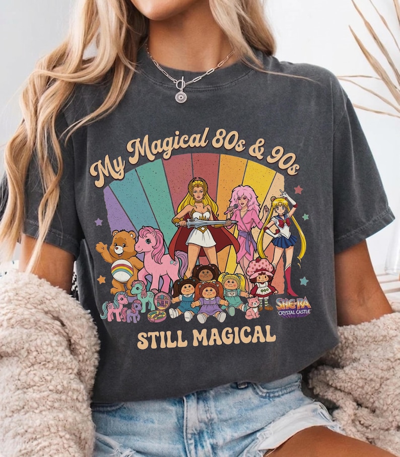 May include: Dark gray t-shirt with a colorful graphic featuring characters from the 80s and 90s, including She-Ra, Sailor Moon, Care Bears, and My Little Pony. The text reads "My Magical 80s & 90s STILL MAGICAL".