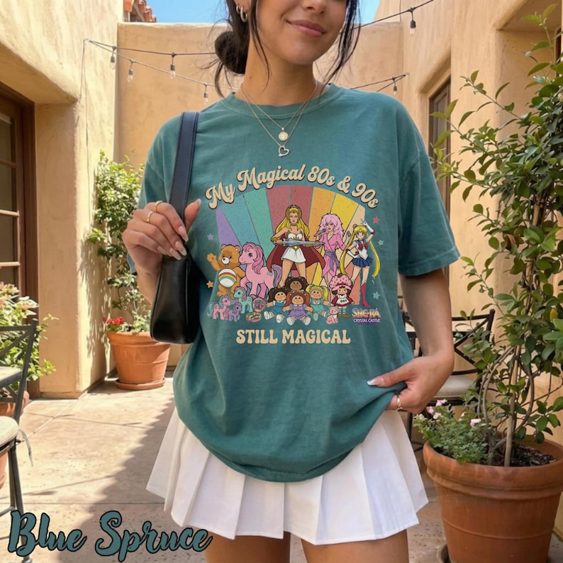 May include: Teal green t-shirt with a colorful graphic featuring cartoon characters and the text "My Magical 80s & 90s STILL MAGICAL". The shirt has a crew neck and short sleeves. The words "Blue Spruce" are at the bottom left.