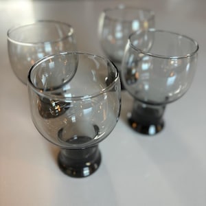 May include: Set of four clear glass drinking glasses with a smoky gray tint. The glasses have a round bowl and a short, thick stem.