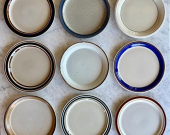 May include: A collection of nine round ceramic plates arranged in a three-by-three grid. Each plate features a distinct rim design, including solid bands of navy, brown, and blue, as well as speckled textures and subtle concentric ring patterns.
