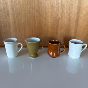 May include: Four ceramic mugs in various colors and styles. The mugs include a white speckled mug with a blue rim, a green pedestal mug, a brown mug, and a white speckled mug with a brown rim. The mugs are on a white surface.