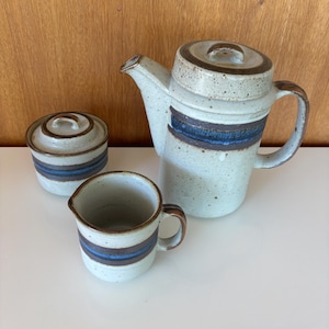 May include: A ceramic tea set with a light blue speckled glaze. The set includes a teapot with a lid, a creamer, and a sugar bowl with a lid. Each piece has a dark blue and brown stripe design.