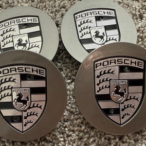 May include: Four silver and gray Porsche wheel center caps. Each cap features the Porsche logo and the word "Porsche" above the Stuttgart emblem, which includes a black horse. The caps are circular and have a metallic finish.