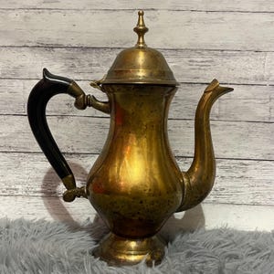 May include: A vintage brass teapot with a black handle and a decorative finial on the lid. The teapot has a curved spout and a bulbous body. The brass has a warm, aged patina, suggesting it is an antique or vintage item.