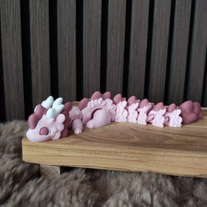 Op de afbeelding: Een roze en bordeauxrood gearticuleerd draakspeelgoed ligt op een houten dienblad. De draak heeft witte hoorns en een gesegmenteerd lichaam. Het speelgoed is gemaakt van een glad materiaal en staat tegen een neutrale achtergrond.