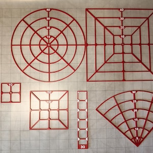 May include: A collection of six red plastic geometric templates featuring grid lines and numerical markings. The set includes a large circle, a large square, two smaller squares, a narrow rectangular strip, and a fan-shaped template.