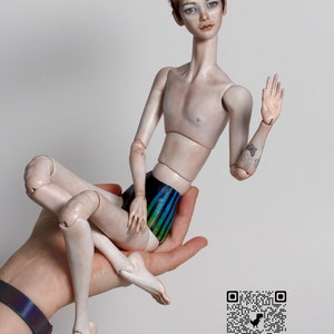 K&ouml;nnte beinhalten: Eine Porzellanpuppe mit kurzen, rotbraunen Haaren, die einen blau-gr&uuml;nen gestreiften Badeanzug tr&auml;gt. Die Puppe sitzt in einer Hand, ein Arm ist zum Gru&szlig; erhoben. Die Puppe hat ein Tattoo auf dem Arm. Das Bild enth&auml;lt einen QR-Code und den Text "dorotesdolls".