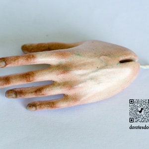 K&ouml;nnte beinhalten: Nahaufnahme einer realistischen Porzellan-Puppenhand mit bemalten Details. Die Hand ist blass mit braunen Akzenten und hat f&uuml;nf Finger. Die Hand liegt flach auf einer wei&szlig;en Oberfl&auml;che.