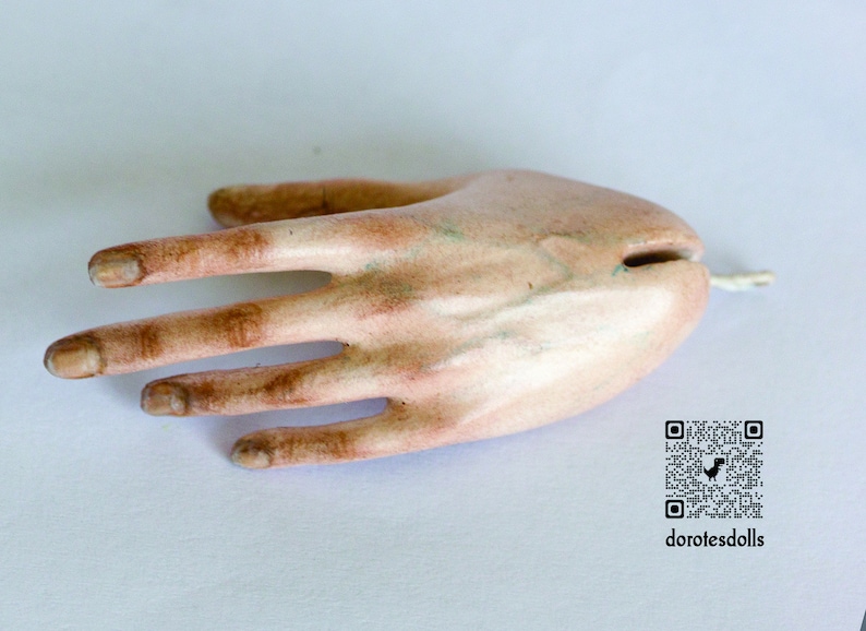 K&ouml;nnte beinhalten: Nahaufnahme einer realistischen Porzellan-Puppenhand mit bemalten Details. Die Hand ist blass mit braunen Akzenten und hat f&uuml;nf Finger. Die Hand liegt flach auf einer wei&szlig;en Oberfl&auml;che.