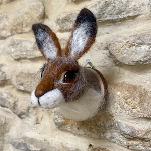 May include: A felted hare head, featuring brown, white, and black wool. The hare has detailed features, including long ears and dark eyes. It is mounted on a stone wall, creating a rustic and natural decorative piece.