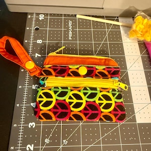 May include: A multi-compartment wallet with a vibrant peace sign pattern in red, orange, yellow, and green. The wallet has a yellow zipper, an orange wrist strap, and a yellow button closure. The background is a black cutting mat.