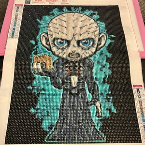 May include: A diamond painting kit featuring a cartoon character with a pale face, blue eyes, and a black robe. The character is holding a small object in their right hand. The background is a dark blue and green color.