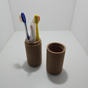 Op de afbeelding: Een bruine, geribbelde tandenborstelhouder met bijpassende deksel. De houder bevat een blauwe en witte tandenborstel, een gele tandenborstel en een tube tandpasta. Ontworpen om mondhygiëneartikelen te beschermen en op te bergen.