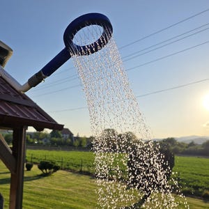 Può includere: Soffione doccia blu per esterni con acqua che cade. Il soffione è circolare con un manico, sullo sfondo di un prato verde, alberi e cielo sereno. Il sole splende luminoso.