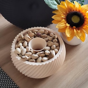 Puede incluir: Un cuenco beige claro con bordes en espiral lleno de pistachos, con una hendidura central. Un girasol amarillo en un jarrón blanco y un cuenco decorativo negro también son visibles sobre una superficie de madera.