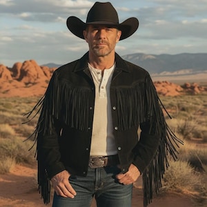 Puede incluir: Un hombre con sombrero de vaquero negro, chaqueta de ante negra con flecos, camisa Henley blanca y vaqueros azules. La chaqueta tiene flecos largos en mangas y pecho. El fondo muestra un paisaje desértico con rocas rojas.
