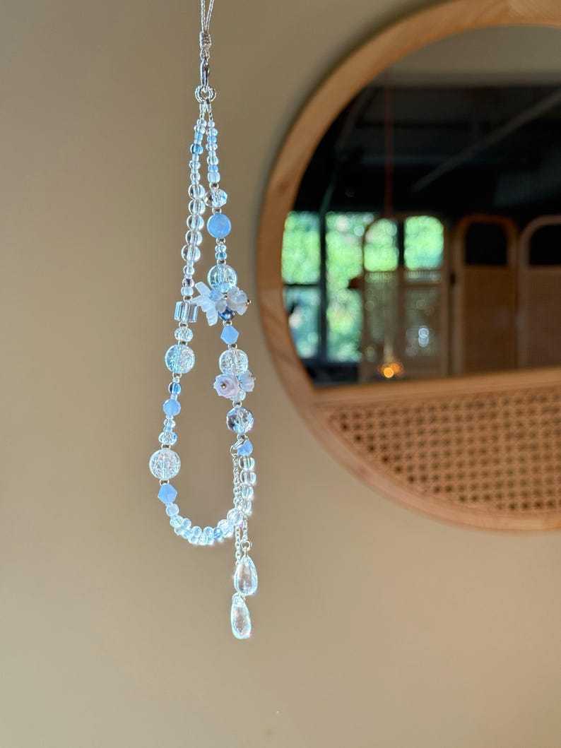 Pu&ograve; includere: Un ornamento di perline fatto a mano con perline trasparenti e azzurro chiaro. Il design include perline rotonde, sfaccettate e a goccia, con un accento floreale. L'ornamento &egrave; appeso a una catena color argento.