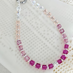 May include: A bracelet featuring a gradient of square-cut crystals transitioning from clear to light pink, then to deep magenta. The beads are separated by small silver-toned spacers, with a single pavé-style bead and a circular silver-toned clasp.