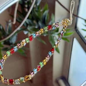 May include: A colorful beaded phone charm with a gold-tone chain. The beads are oval-shaped and in shades of red, yellow, green, blue, and purple. The charm is displayed on a reflective surface.