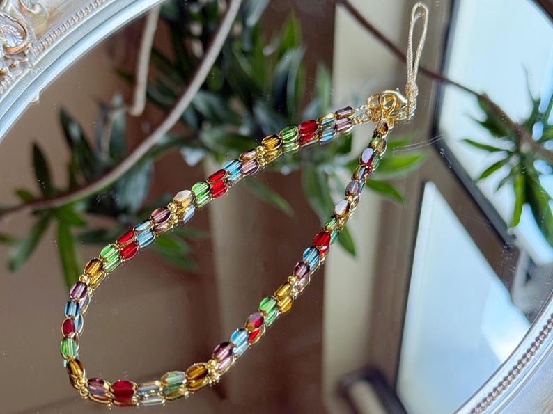May include: A colorful beaded phone charm with a gold-tone chain. The beads are oval-shaped and in shades of red, yellow, green, blue, and purple. The charm is displayed on a reflective surface.