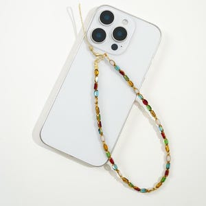 Peut inclure: Un smartphone blanc avec une lanière de téléphone à perles colorées. La lanière est composée d'une chaîne dorée ornée de perles rouges, vertes, bleues et jaunes. Le téléphone possède trois objectifs.